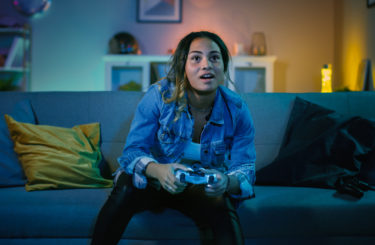 Beautiful Excited Young Black Gamer Girl Sitting on a Couch and Playing Video Games on a Console. She Plays with a Wireless Controller. Cozy Room is Lit with Warm and Neon Light.