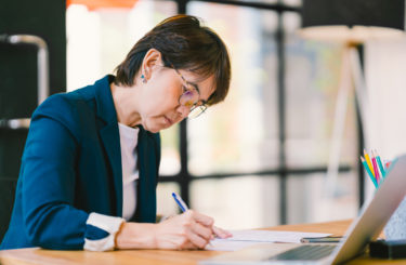 Beautiful middle age Asian woman working on paperwork in modern contemporary office, with laptop computer. Business owner, entrepreneur, executive manager, or employee office worker concept