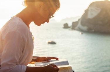 Young woman reading book at sunset in front of the sea on Ponza island coast, sitting on a wall with view of the ocean.