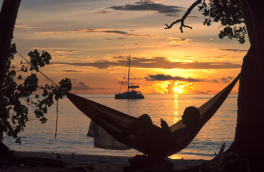 Beach vacations, silhouette of a man reading in hammock at sunset on tropical island.