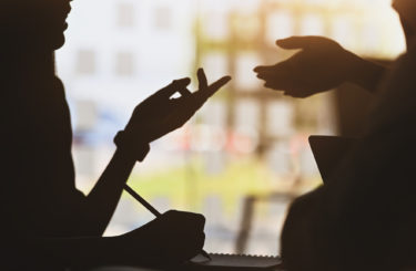 silhouette of young designer team standing with a white blank screen laptop and notebook in hands while discussing/talking about them new project with the modern office as background.