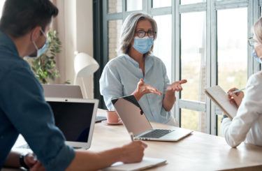 Old female mentor coach wearing face mask training young workers at group meeting in reopen office. Middle aged businesswoman teacher working with students at university class. Social distance concept