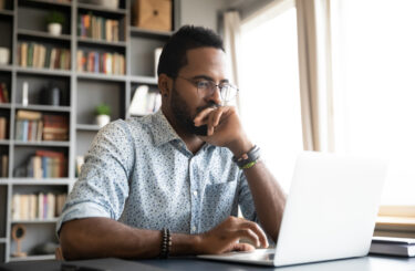 Focused concentrated young african businessman sit at desk look at laptop, serious afro american male professional analyst working online on computer data watch webinar thinking of problem solution