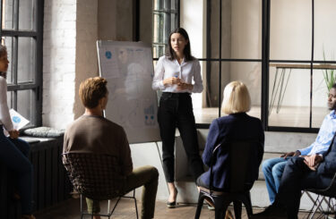 Confident lady business trainer coach leader give flip chart presentation consulting clients teaching employees training team people speaking explaining strategy at marketing workshop concept