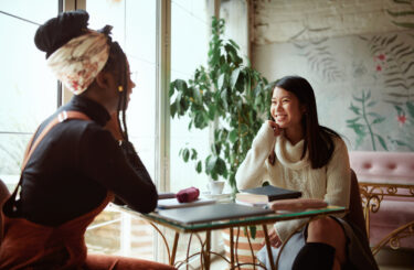 Two happy multicultural female college girls sit in the cafeteria, chatting and gossiping. Female students and girls talk.