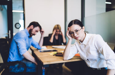 Exhausted female employee in business style clothing and glasses working with coworkers at desk and leaning on hand seeking solution of problem at office and looking at camera