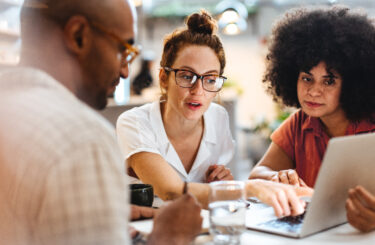 Business women having a work lunch in a café, exchanging ideas and discussing their projects with a client. Young business team using a laptop as they sit around a coffee table.